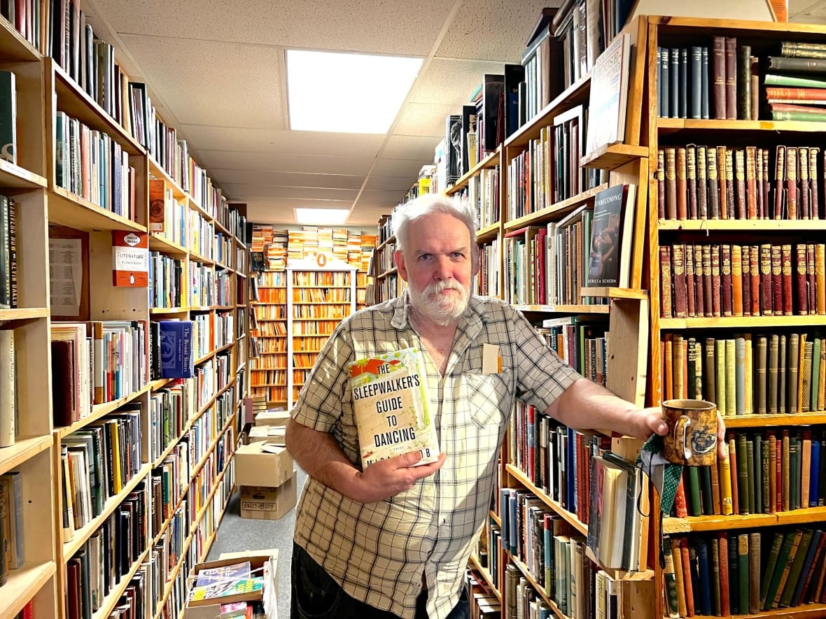 A man with white hair and a beard holds a book inside a crowded bookstore. He stands in between two shelves stacked with books. 