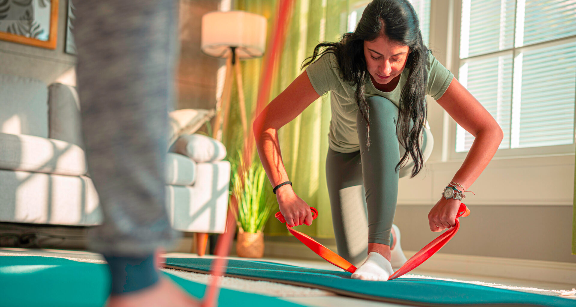 Woman in sportswear kneels down and holds both ends of a long resistance band, her front foot is on the middle of the resistance band. She is in a domestic setting with a couch in the background and someone's lower leg in the foreground