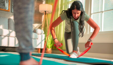 Woman in sportswear kneels down and holds both ends of a long resistance band, her front foot is on the middle of the resistance band. She is in a domestic setting with a couch in the background and someone's lower leg in the foreground