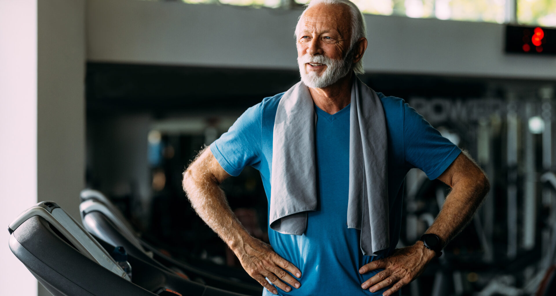 A man stands on a treadmill in a gym, with his hands on hips and a sweat towel draped over his shoulders.