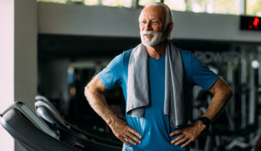 A man stands on a treadmill in a gym, with his hands on hips and a sweat towel draped over his shoulders.