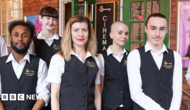 James Jervis sat in a seat in a cinema with black and gold chairs velvet chairs. He is wearing a white shirt and has thick brown hair and is smiling at the camera.