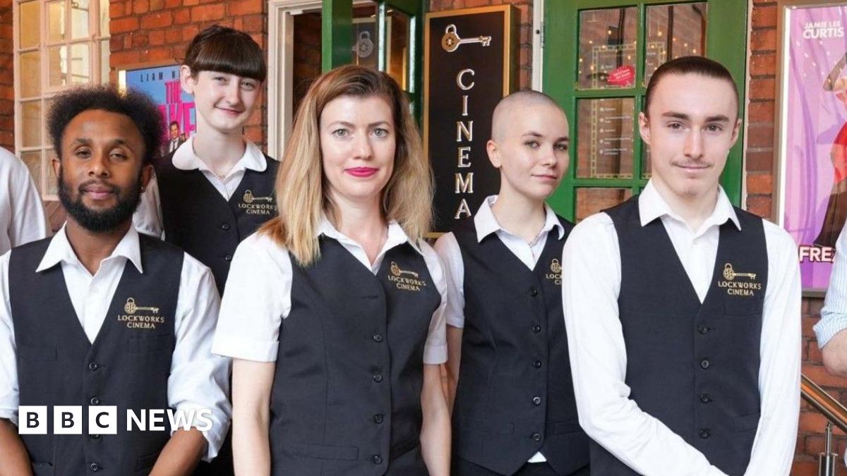 James Jervis sat in a seat in a cinema with black and gold chairs velvet chairs. He is wearing a white shirt and has thick brown hair and is smiling at the camera.