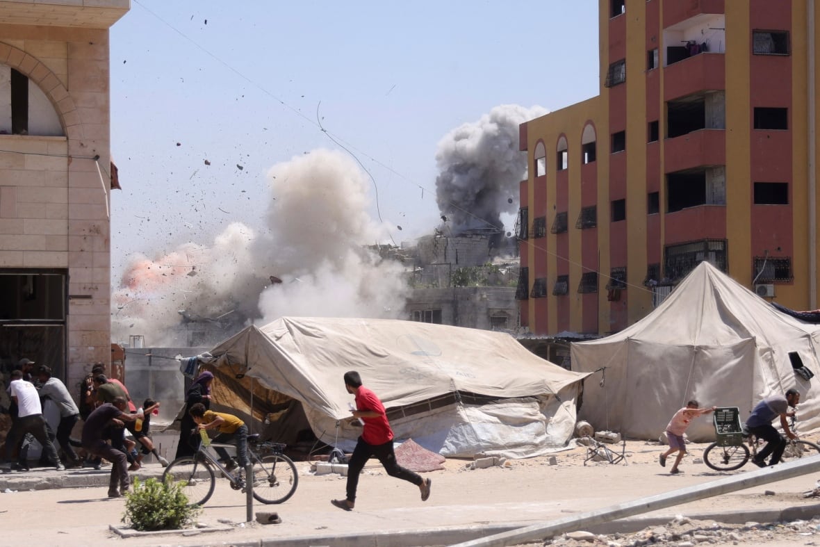 Palestinians on a sidewalk near a building rushing for cover as an airstrike hits a building nearby in the distance.