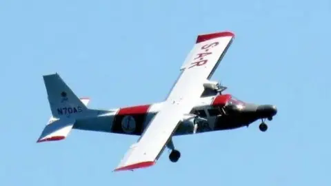 Channel Islands Air Search The aircraft, called the Lions’ Pride, airborne. It is white with red writing on the right wing which says 'S-A-R' and has a red block in the middle. The wing at the back has red text on it. The the front there are two windows on the left and is grey.