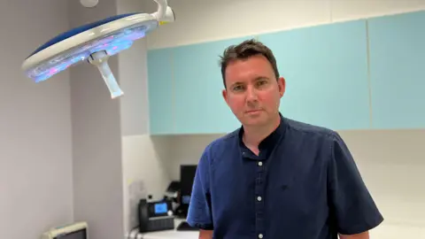 Photo of a man with short brown hair looking at the camera. He is wearing a navy blue button-down shirt. He is standing in a doctor's examination room.