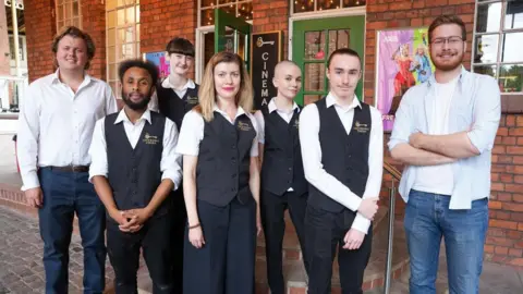 City of Wolverhampton Council Seven people are standing outside a building with two green doors, one of which is open. Everyone, except the men on the far left and far right, are wearing black-and-white Lockworks Cinema uniforms that include its name.