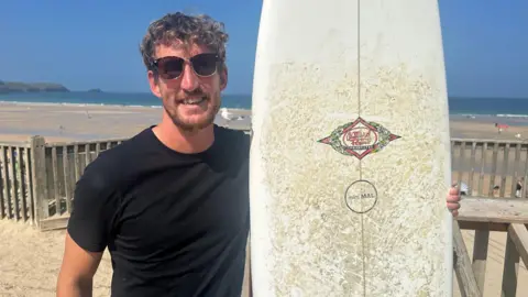 A man in a dark T-shirt and glasses holding a surfboard with a wooden fence and sea behind him.