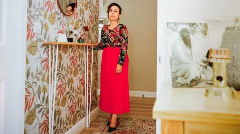Mana Baoosh A lady wearing a red and black floral top and a long red skirt. She has her hand leaning on a shelf looking at the camera and in the foreground an old black and white photo of someone can be seen sat on a mantle piece.