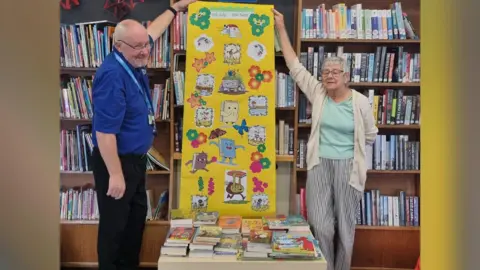 Friends of Woodston Library Volunteers - a man wearing a blue T-shirt and black trousers and a woman wearing a mint coloured T-shirt, a white cardigan and striped trousers, holding a yellow poster placed behind a table of books 