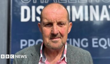 Cliff Donaldson is looking straight into the camera with a neutral expression on his face. He is an older man with a bald head, and is wearing a grey plaid blazer and a red and white flannel shirt underneath. He is standing outside the Equalty Commission building's blue signs.