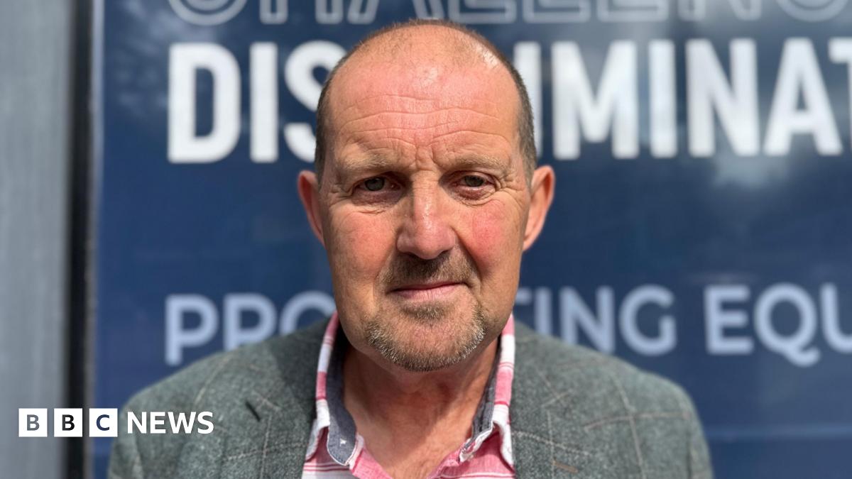 Cliff Donaldson is looking straight into the camera with a neutral expression on his face. He is an older man with a bald head, and is wearing a grey plaid blazer and a red and white flannel shirt underneath. He is standing outside the Equalty Commission building's blue signs.