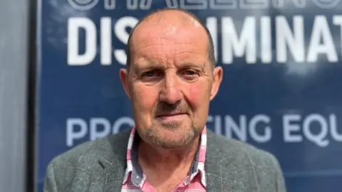 BBC Cliff Donaldson is looking straight into the camera with a neutral expression on his face. He is an older man with a bald head, and is wearing a grey plaid blazer and a red and white flannel shirt underneath. He is standing outside the Equalty Commission building's blue signs.