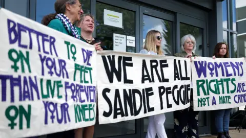 Getty Images Several women hold banners supporting gender critical nurse Sandie Peggie - the banners saying We Are All Sandie Peggie, better to die on your feet than live forever on your knees and women's rights are not a hate crime