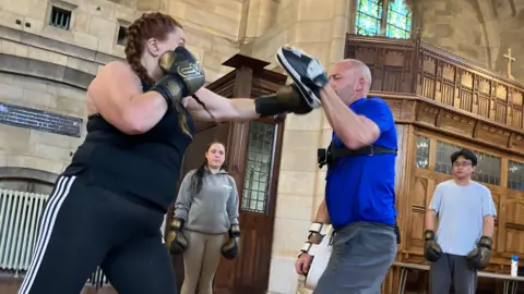Ben Cookson takes a punch from a woman in black exercise clothes while wearing a pad on his left hand during a boxing class in a church. Two other people wearing boxing gloves are watching the action.