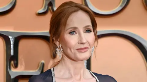 Getty Images Head shot of Harry Potter author JK Rowling, She has red hair, tied back in a ponytail and is wearing long diamond earrings. She is wearing a black dress, with a diamond trim. Rowling is standing in front of a pink backdrop advertising "Fantastic Beasts: The Secret of Dumbledore". She is looking off to the left of the camera.
