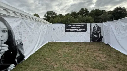 White sheets erected in a field showing a tribute to Ozzy Osbourne. Two black and white photos of him are on the sheets alongside a black text box saying 'John "Ozzy" Osbourne/ 1948 - 2025'. The white sheets are covered with hand-written tributes.