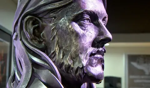 BBC/Kevin Reide A close-up in profile of a metal bust of Lemmy from Motorhead, with purple light shining on it. The bust is silver in colour and has long hair and a beard.