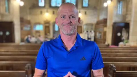 Ben Cookson smiles while stood between two rows of church pews. He has a blue Adidas t-shirt on.