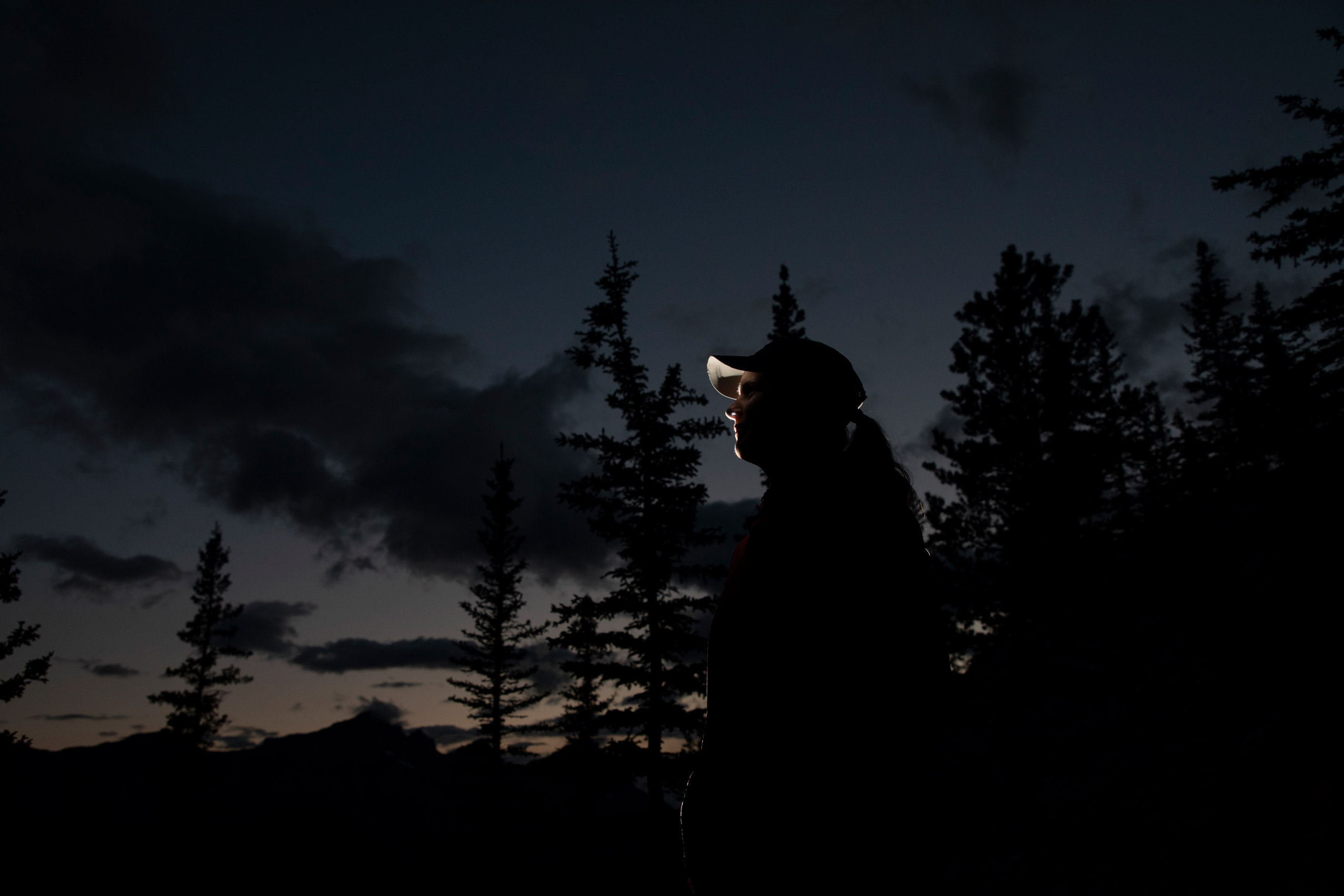 A side profile of a woman in a baseball cap in front of the silhouettes of trees in the dark