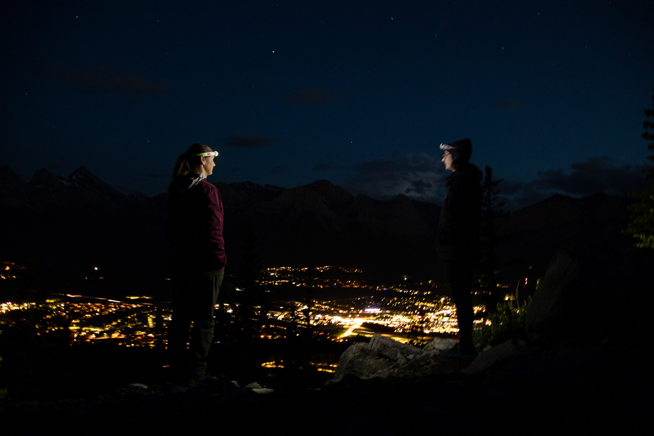 Two hikers wearing headlamps look out over a valley lit up by Canmore lights, with stars visible above