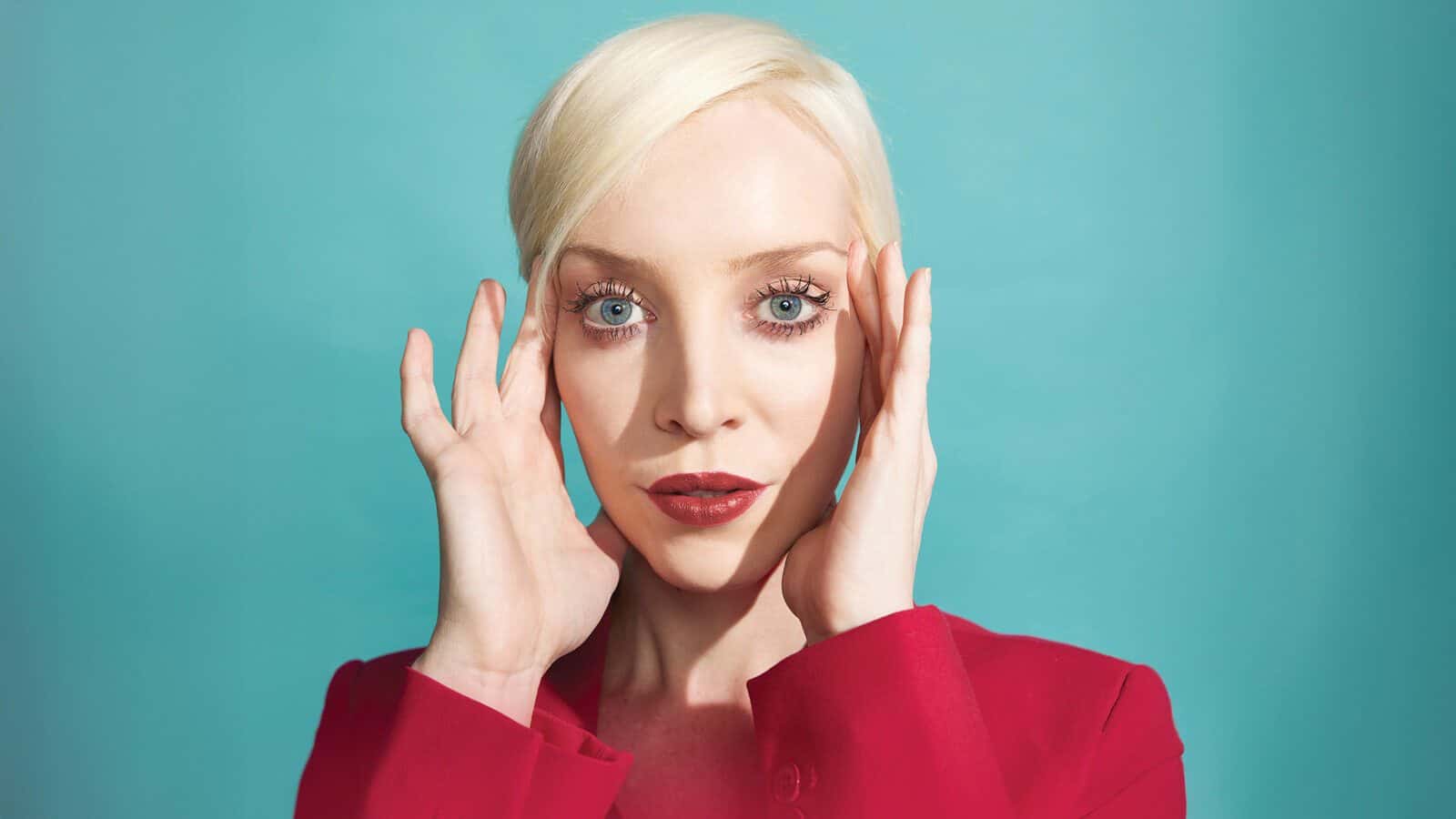 A person with short platinum blonde hair in a red jacket poses against a turquoise background, channeling Kira Jane Buxton’s bold style, touching their head with both hands and looking directly at the camera.