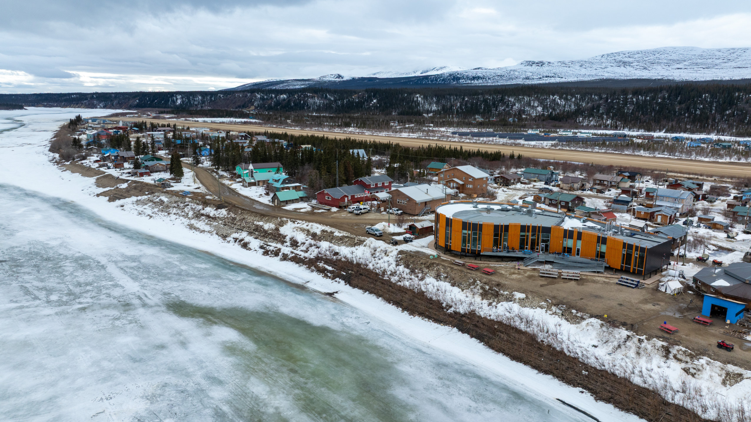 This aerial photo depicts Old Crow, Yukon, an Arctic community on the shore of the Porcupine River. It's spring, and while most of the snow has melted, the river is still frozen. The Old Crow Community Centre is in the photo's foreground.
