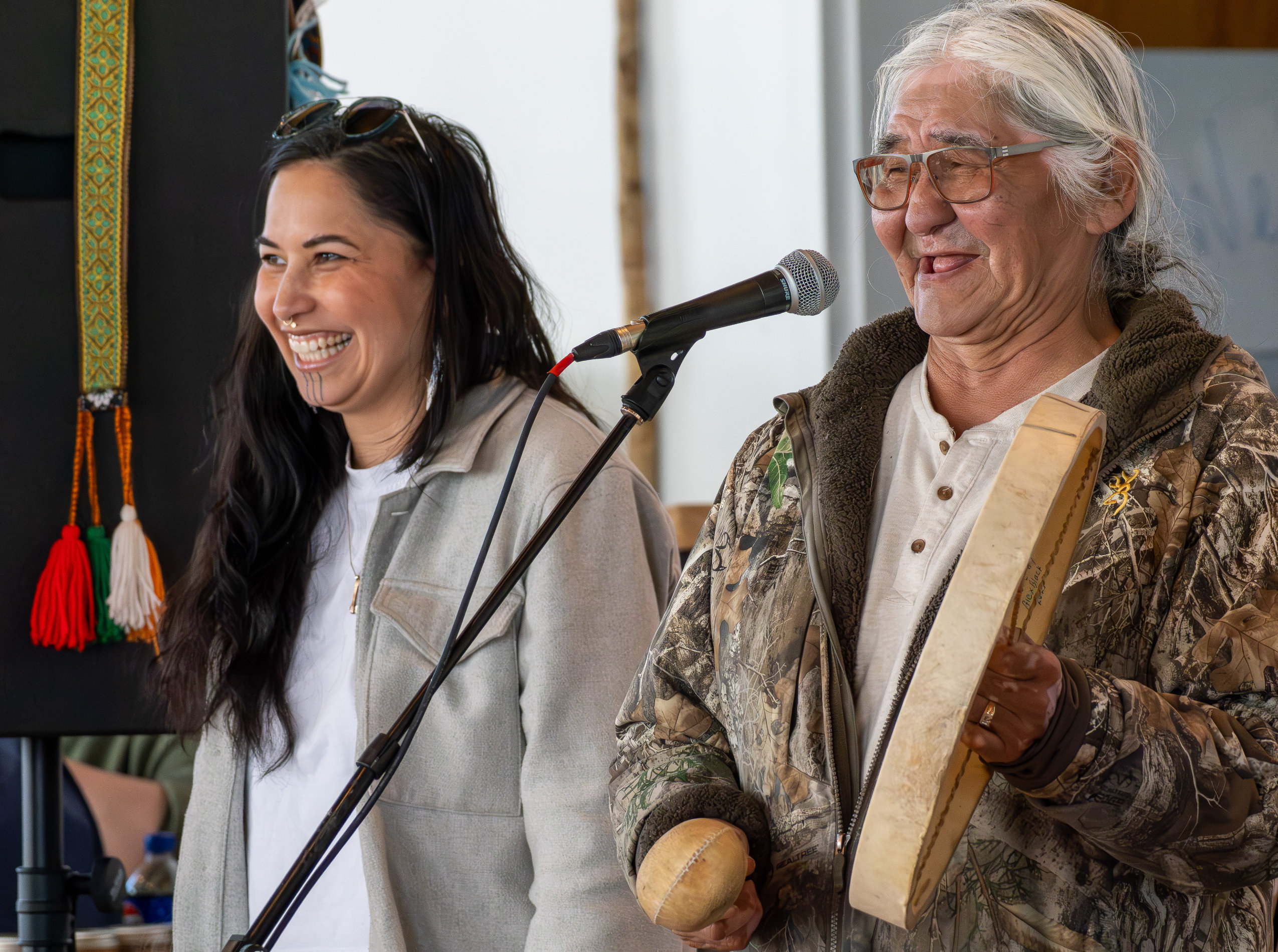 Two people stand smiling and looking toward the left. A microphone is positioned in front of the person on the right, and he holds a hand drum.
