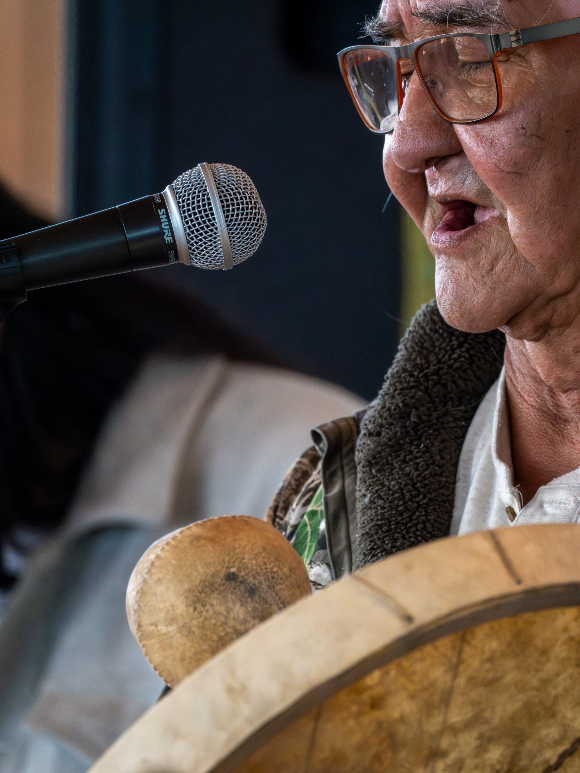 A close-up image of a man singing into a microphone and holding a drum.