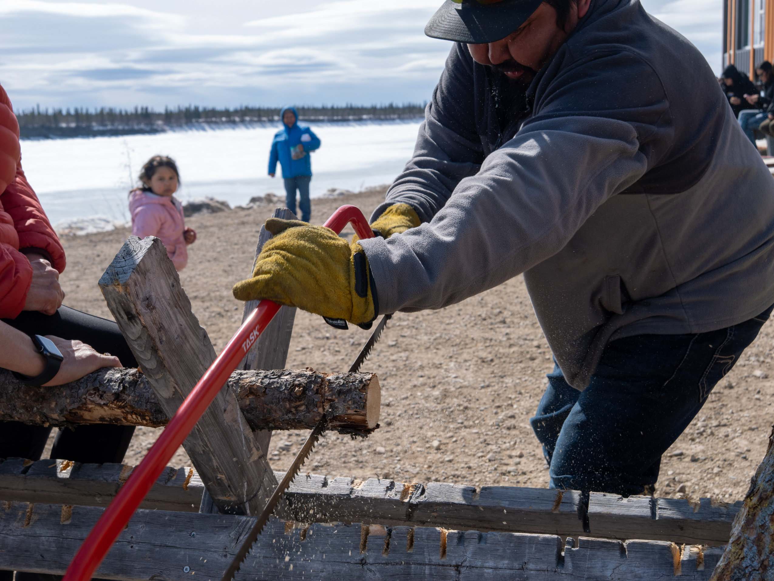 A man saws through a small tree branch as children observe in the background.