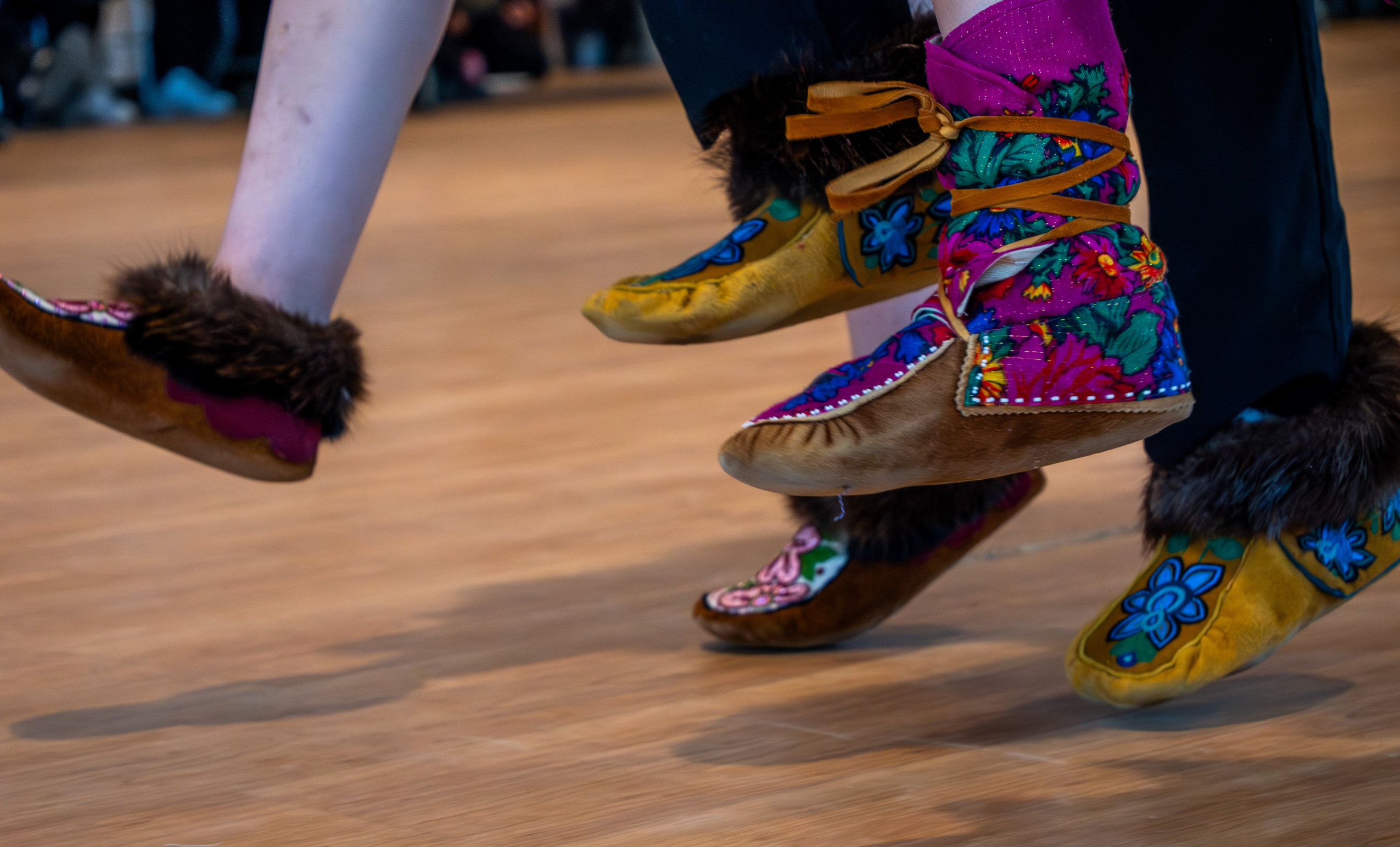 Several feet wearing decorative moccasins jump off the ground during a performance of a traditional dance.