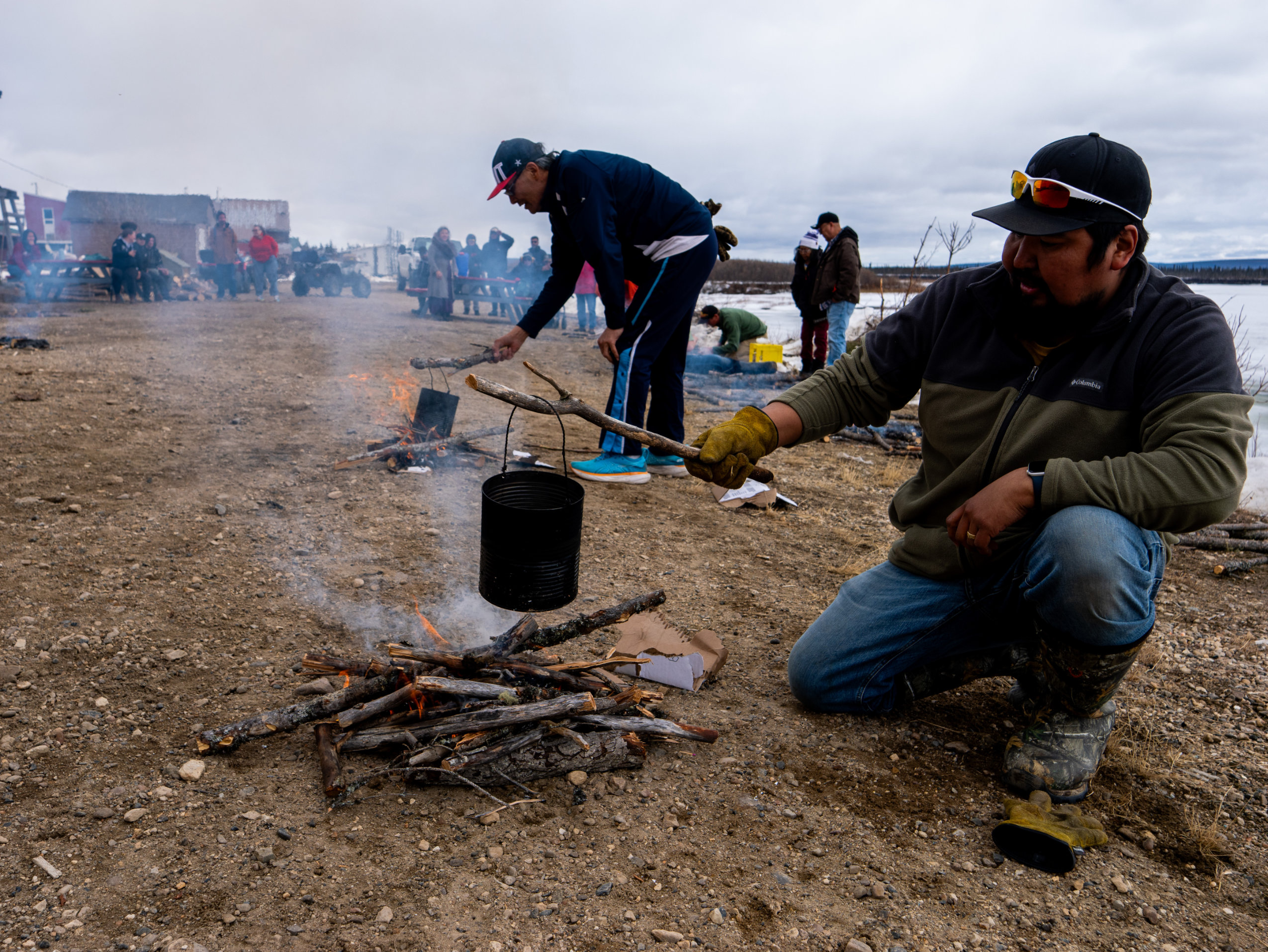 Two men suspend blackened cans full of water above bonfires to boil tea.