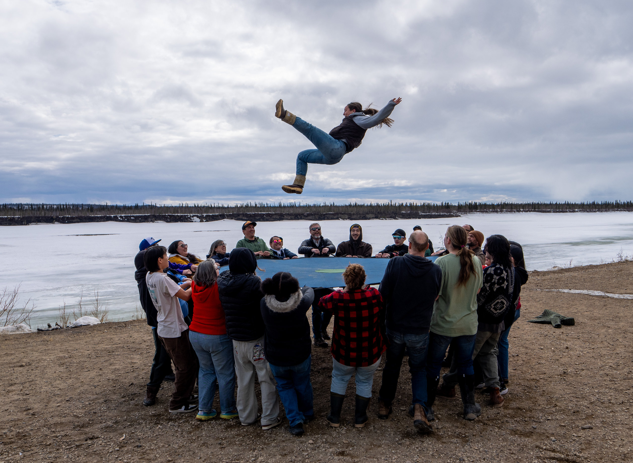 About two-dozen people hold a circular trampoline and propel a person into the air. In the background, a frozen river.