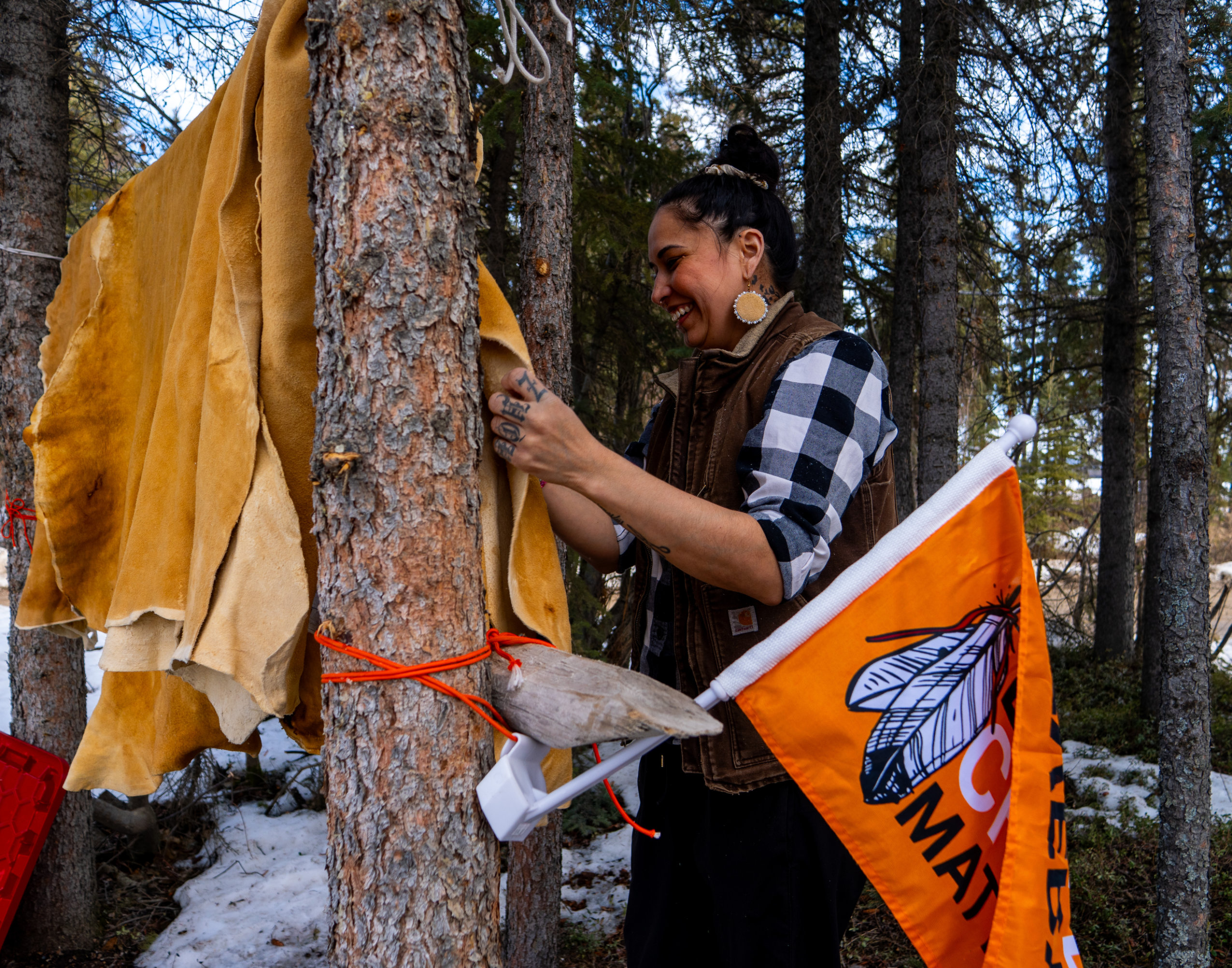 A young woman smiles as she handles a dried caribou skin that his hanging from some trees.