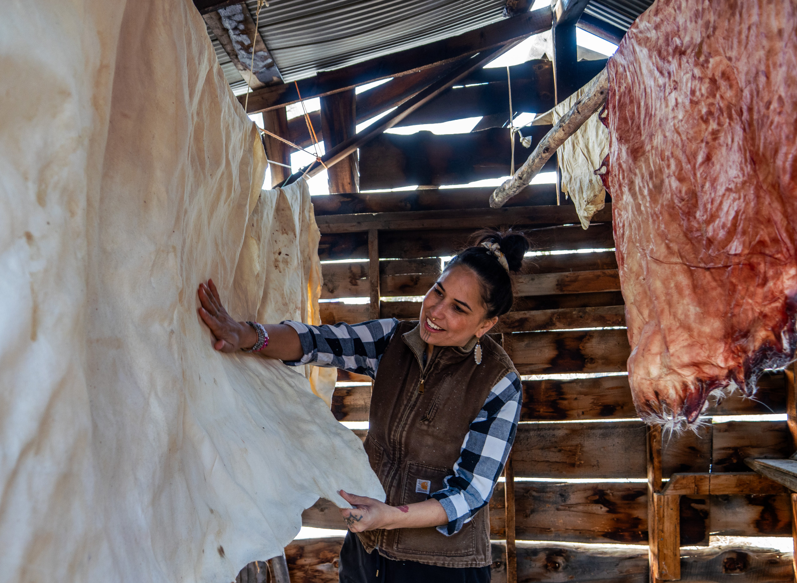 A woman shows off her caribou hides, which are hanging in a wooden structure.