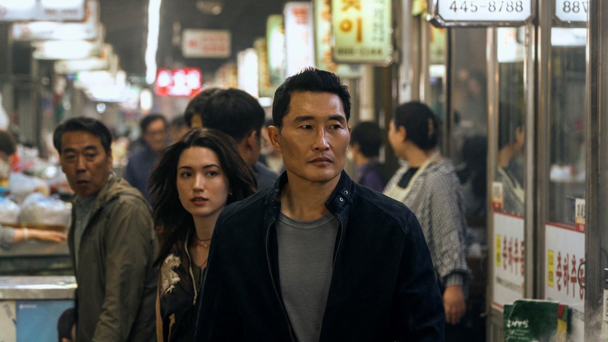 David leads Rebecca through a crowded Korean market. He wears a black jacket over a gray shirt. 