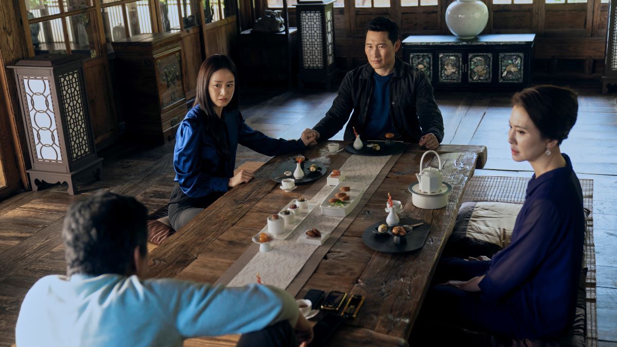 Eunju and David hold hands at one end of a dining table, facing Eunju's parents, seated opposite them. 