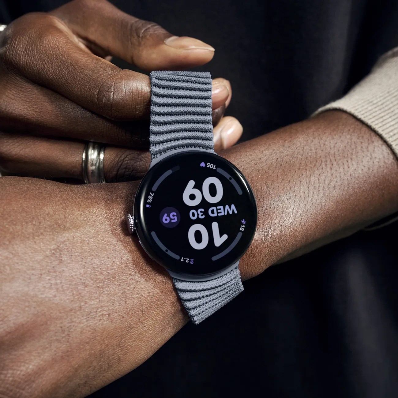 A close-up of a person wearing a smartwatch with a round black face and a textured gray fabric strap. The watch display shows the time as 10:09, the date as Wednesday the 30th, and various health metrics including heart rate and activity progress. The person has dark skin and is adjusting the watch with their other hand, which has a silver ring on the ring finger.