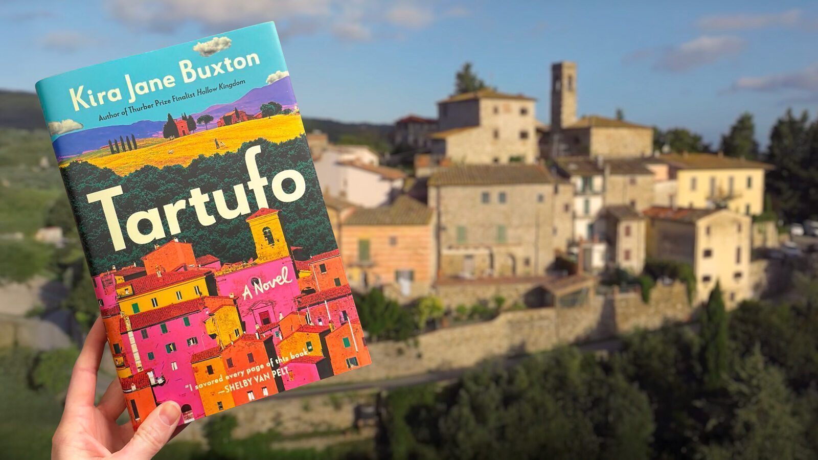 A hand holds "Tartufo" by Kira Jane Buxton before a sunlit, rustic Italian village with stone buildings and a tower.