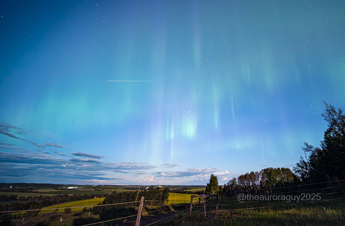 Meteors are visible streaking through a dark blue sky wreathed in a green aurora above a rural landscape.