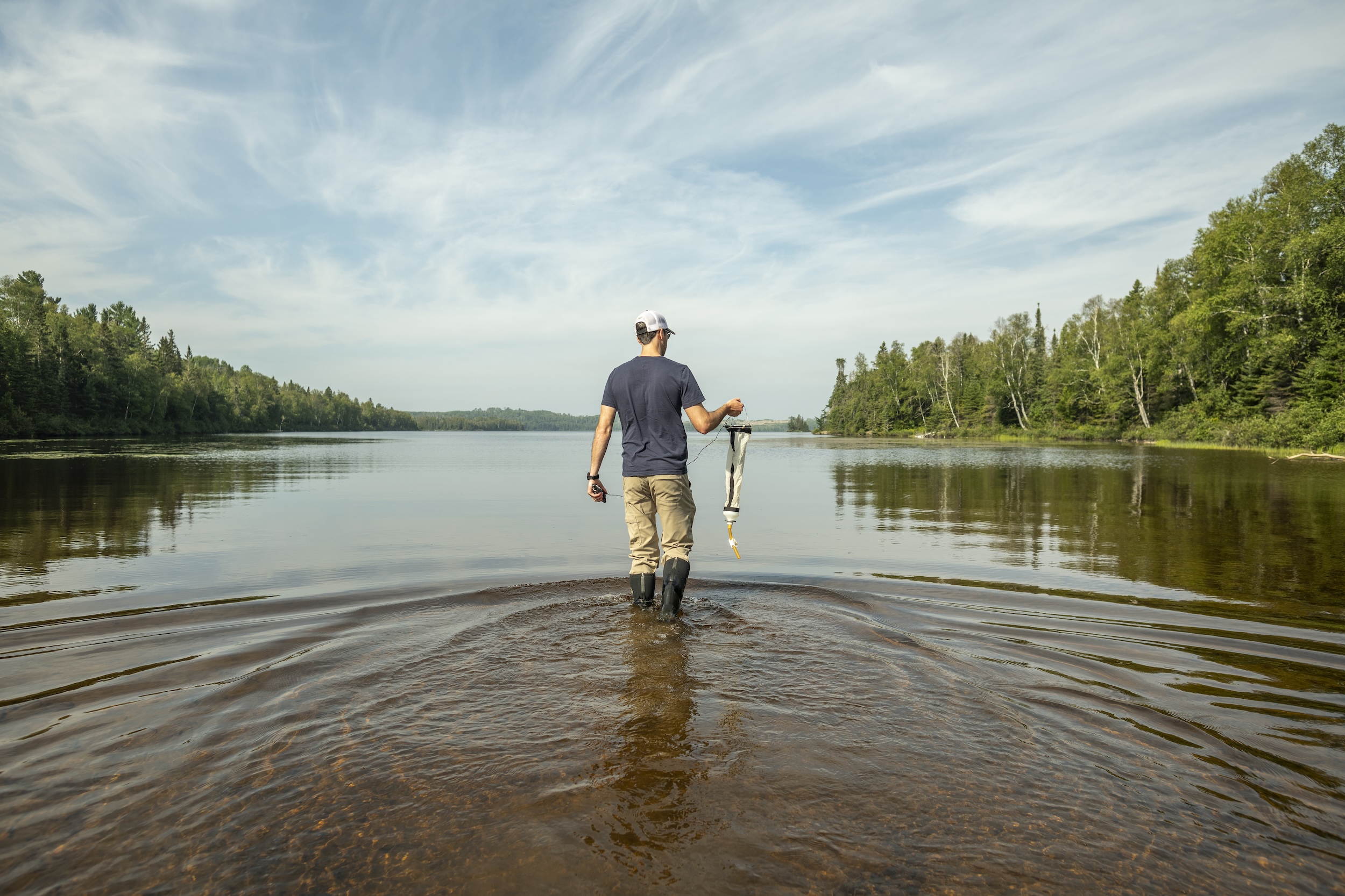 A man walks into shallow water holding a plankton net, with blue skies above