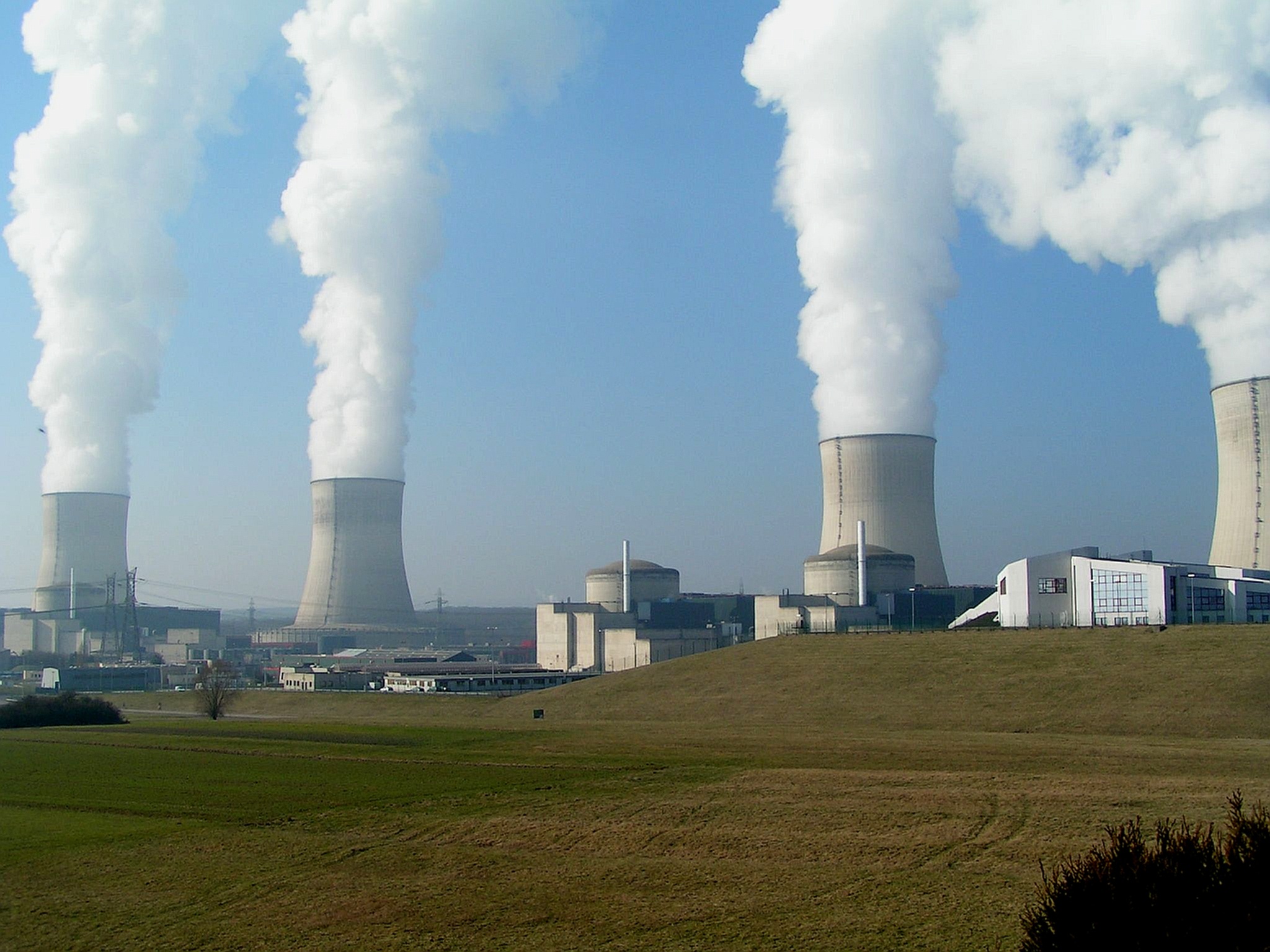 A nuclear power plant with four large cooling towers emitting white steam, set against a blue sky with grassy fields in the foreground.