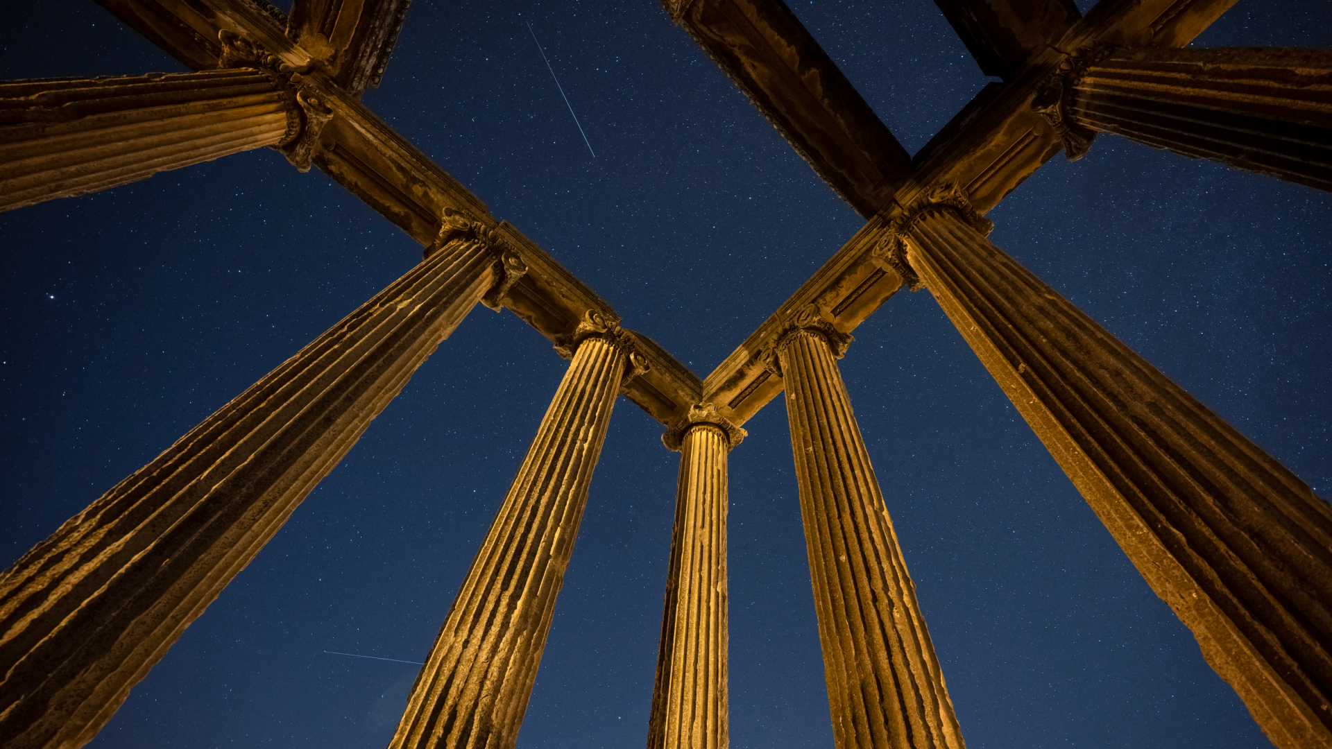A picture facing up through the columns of a ruined temple, which is softly illuminated by light. Meteors are visible streaking through the night sky beyond.