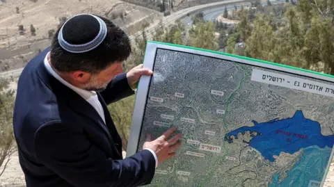 Reuters Israeli Finance Minister Bezalel Smotrich and a woman hold a map that shows the long-frozen E1 settlement scheme, that would split East Jerusalem from the occupied West Bank, on the day of a press conference near the Israeli settlement of Maale Adumim, in the Israeli-occupied West Bank, August 14, 2025.