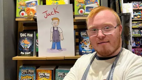 Supplied Jack Dillon smiles at the camera next to an illustration of himself from the book. He has short ginger hair and a beard, blue eyes and is wearing glasses, a white shirt and a blue apron. 