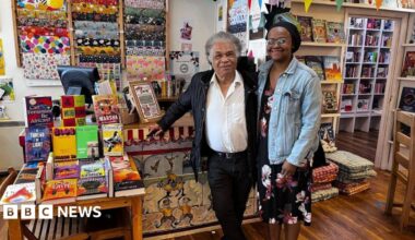 A man and a woman are stood in the middle, in front of a cashier desk in a bookshop. The man is on the left and has grey hair. He is wearing a white shirt, unbuttoned at the collar, and a black jacket, with black trousers and a belt. He is leaning against the desk with his right arm. The woman on the right is wearing a black hair bonnet, and is wearing a black dress with red and white floral decorations and a light blue denim jacket. To their left, there is a table displaying a colourful selection of books. There is a computer and more books on the desk, and behind the desk there is a display of wrapping paper hung on the white wall. There are more book shelves behind the two people, on their right.