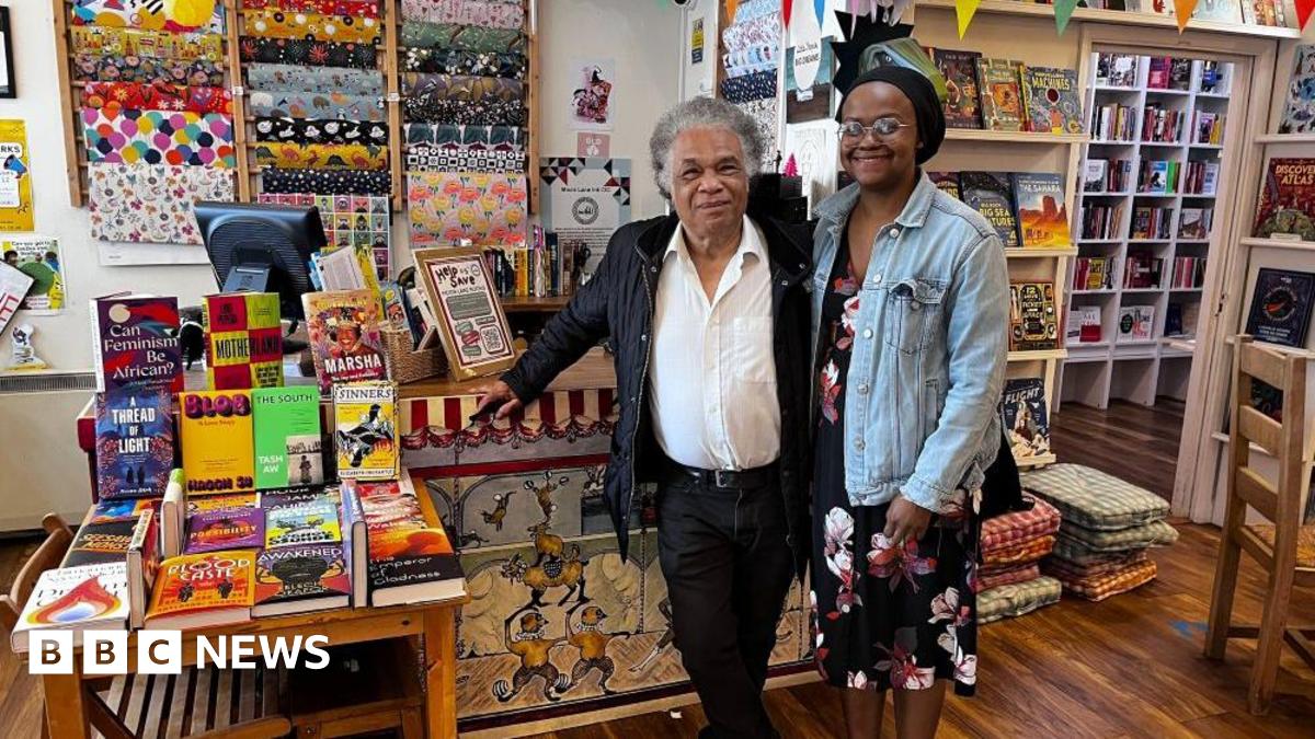 A man and a woman are stood in the middle, in front of a cashier desk in a bookshop. The man is on the left and has grey hair. He is wearing a white shirt, unbuttoned at the collar, and a black jacket, with black trousers and a belt. He is leaning against the desk with his right arm. The woman on the right is wearing a black hair bonnet, and is wearing a black dress with red and white floral decorations and a light blue denim jacket. To their left, there is a table displaying a colourful selection of books. There is a computer and more books on the desk, and behind the desk there is a display of wrapping paper hung on the white wall. There are more book shelves behind the two people, on their right.
