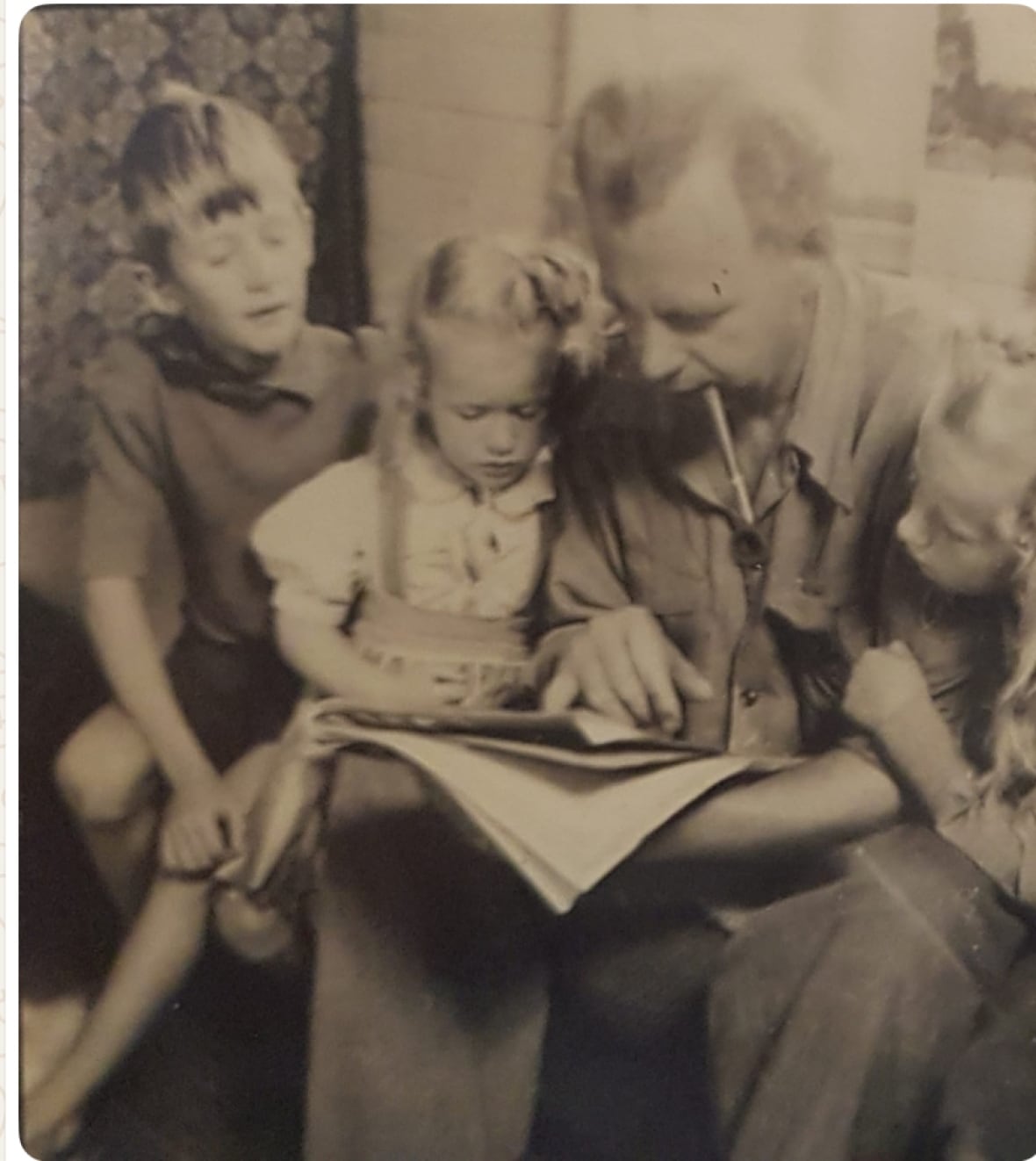 A black and white photo of a man reading a book to three children, one boy and two girls.
