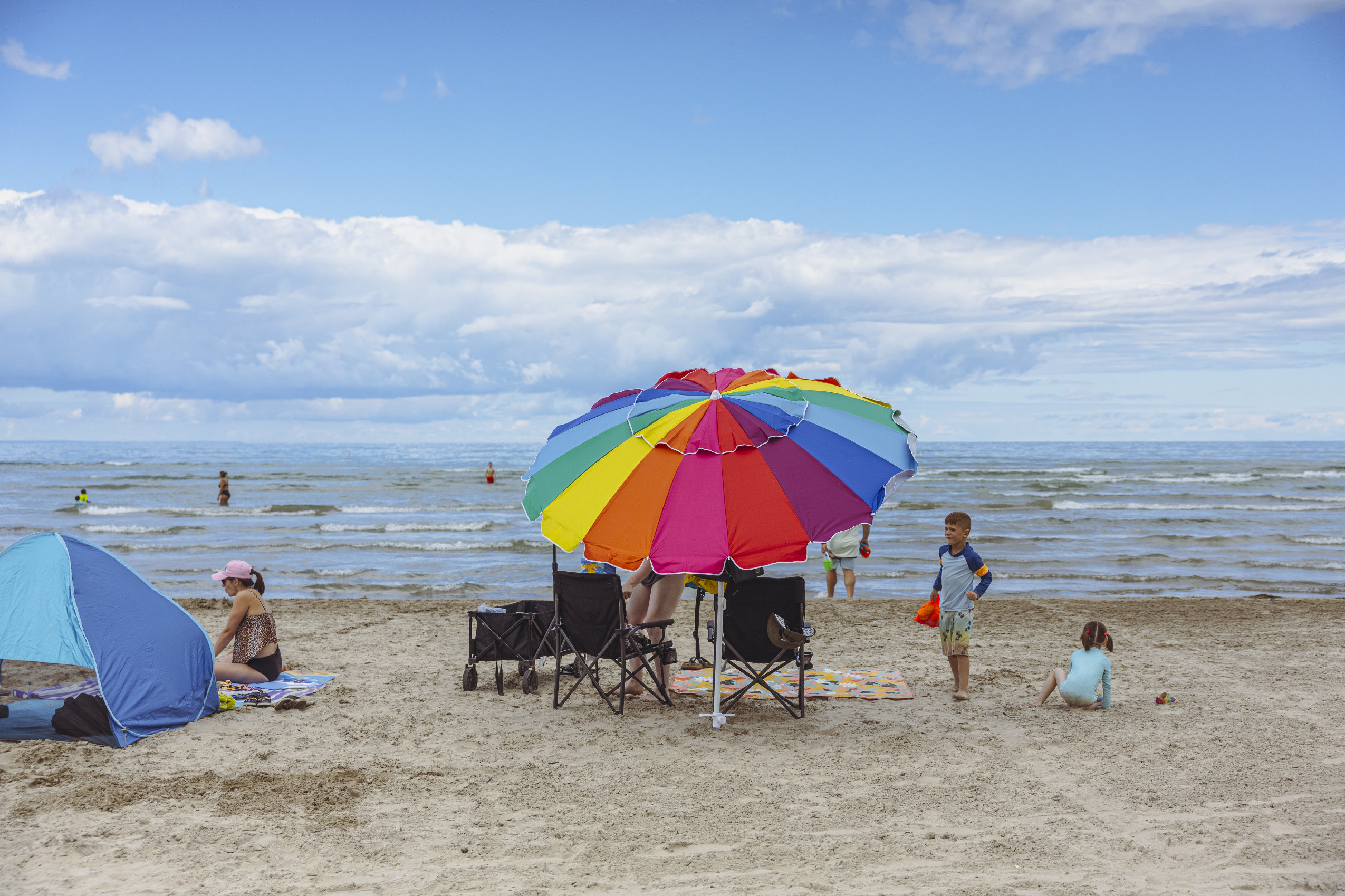 Bathers are seen swimming and hanging out on the sand at Wasaga Beach. A big, multi-coloured beach umbrella hides the heads of some of the beachgoers.