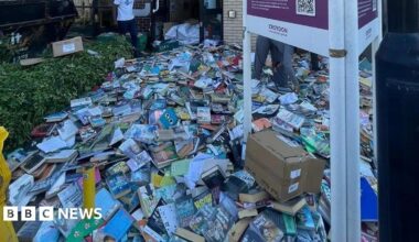 Piles of books cover the pavement outside Broad Green Library in Croydon, many in good condition and some in open boxes. Two people are visible among the stacks, with a black skip to the left and a library sign in the foreground.